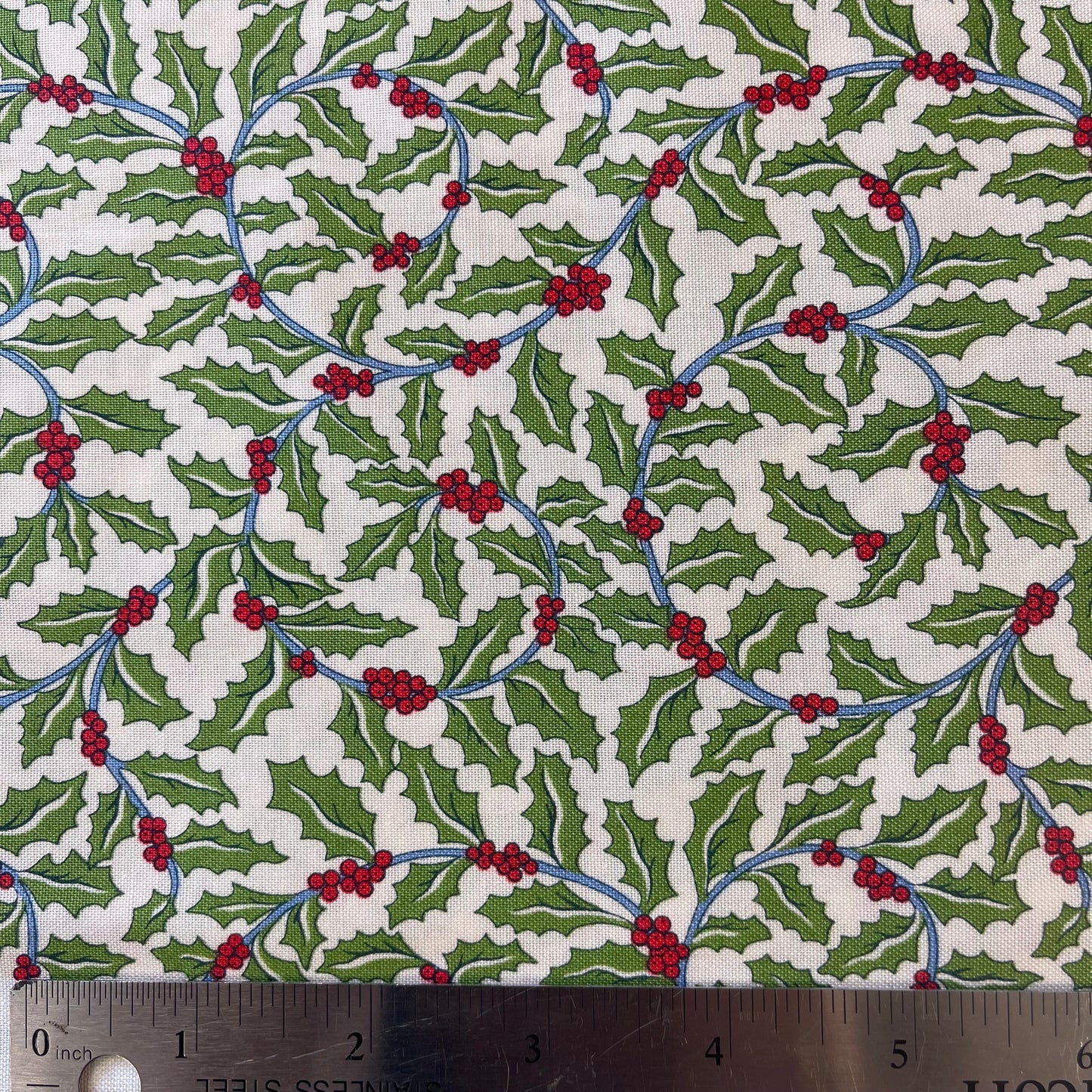 A close-up of patterned fabric with a repeating holly design featuring green leaves, red berries, and blue stems on an off-white background from Liberty of London Fabrics for quilting and crafting.