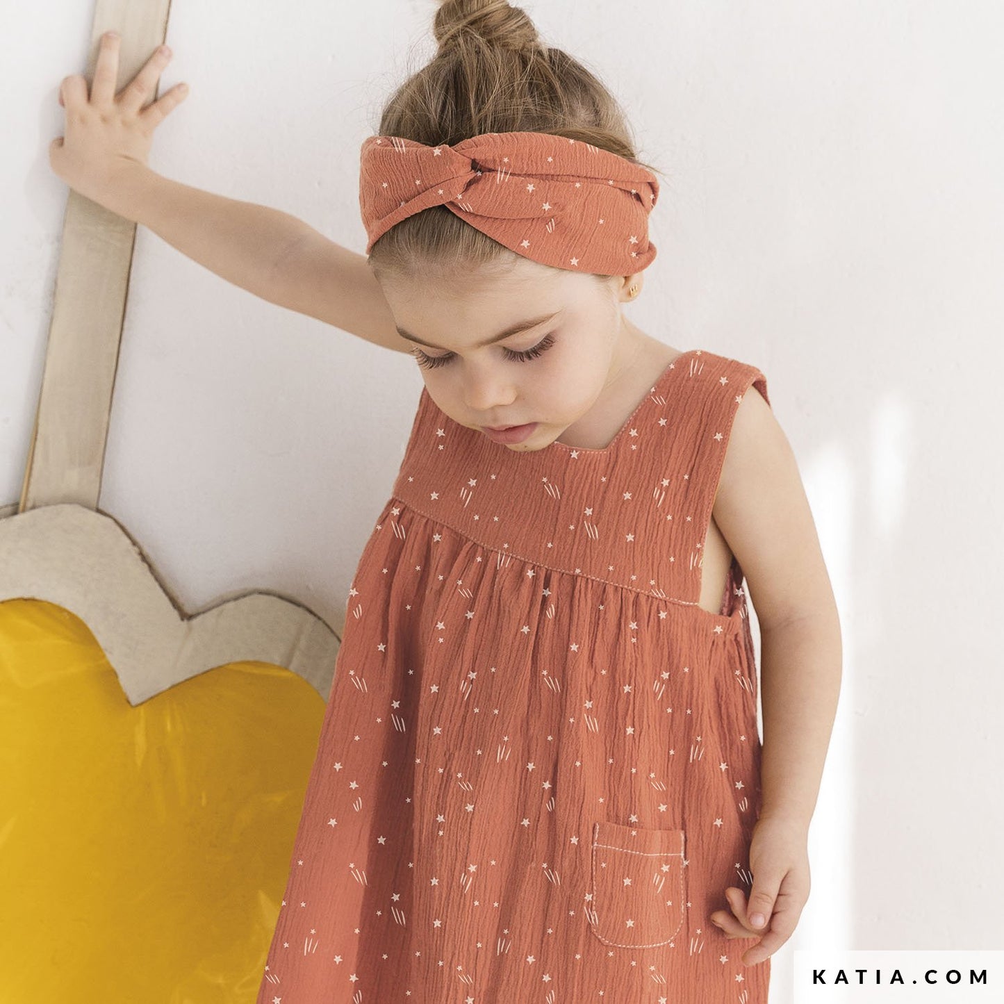 Young girl wearing a coral colored dress with a matching headband, standing against a white wall.