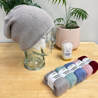 Gray knit beanie on a mannequin head, yarn skeins in various colors with labels, and a plant in the background.