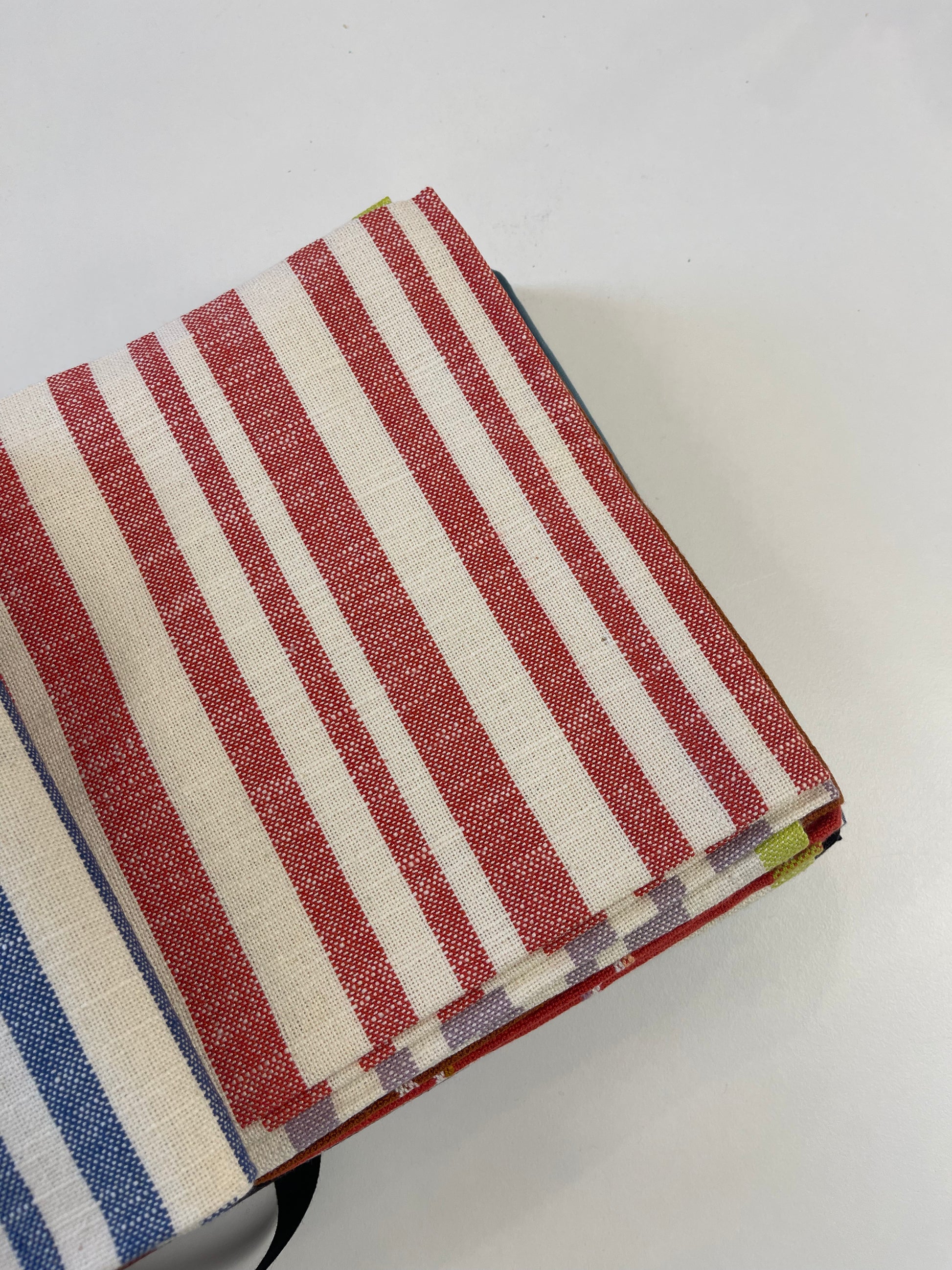 A folded stack of striped woven fabrics on a plain white background, featuring red and off-white stripes with a blue and white stripe pattern partially visible at the bottom left.