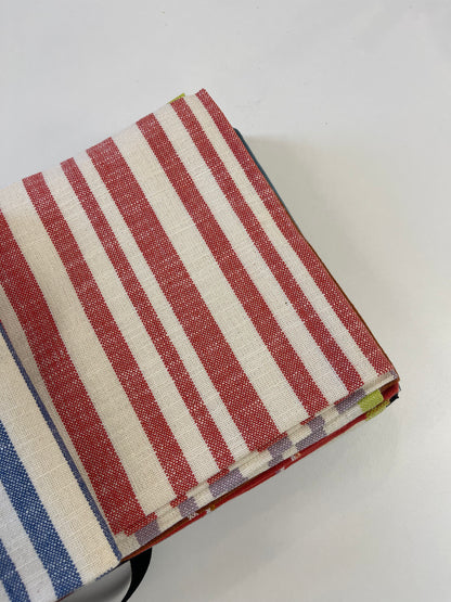 A folded stack of striped woven fabrics on a plain white background, featuring red and off-white stripes with a blue and white stripe pattern partially visible at the bottom left.