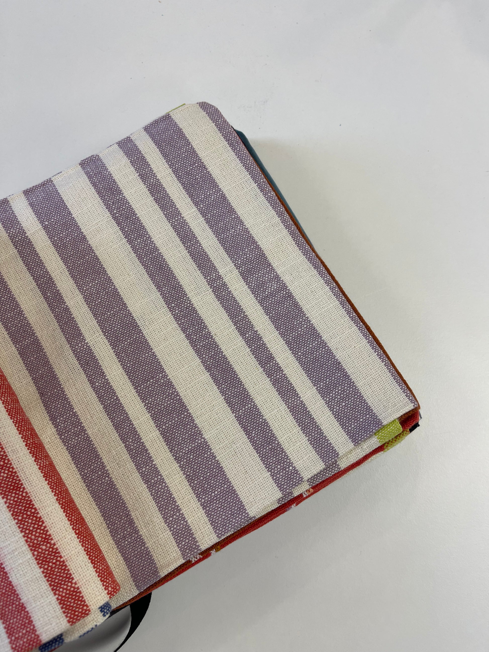 A close-up of a striped fabric item, likely a notebook cover or pouch, featuring vertical stripes in lavender, cream, and red, with a black strap visible at the bottom left, set against a plain white background.