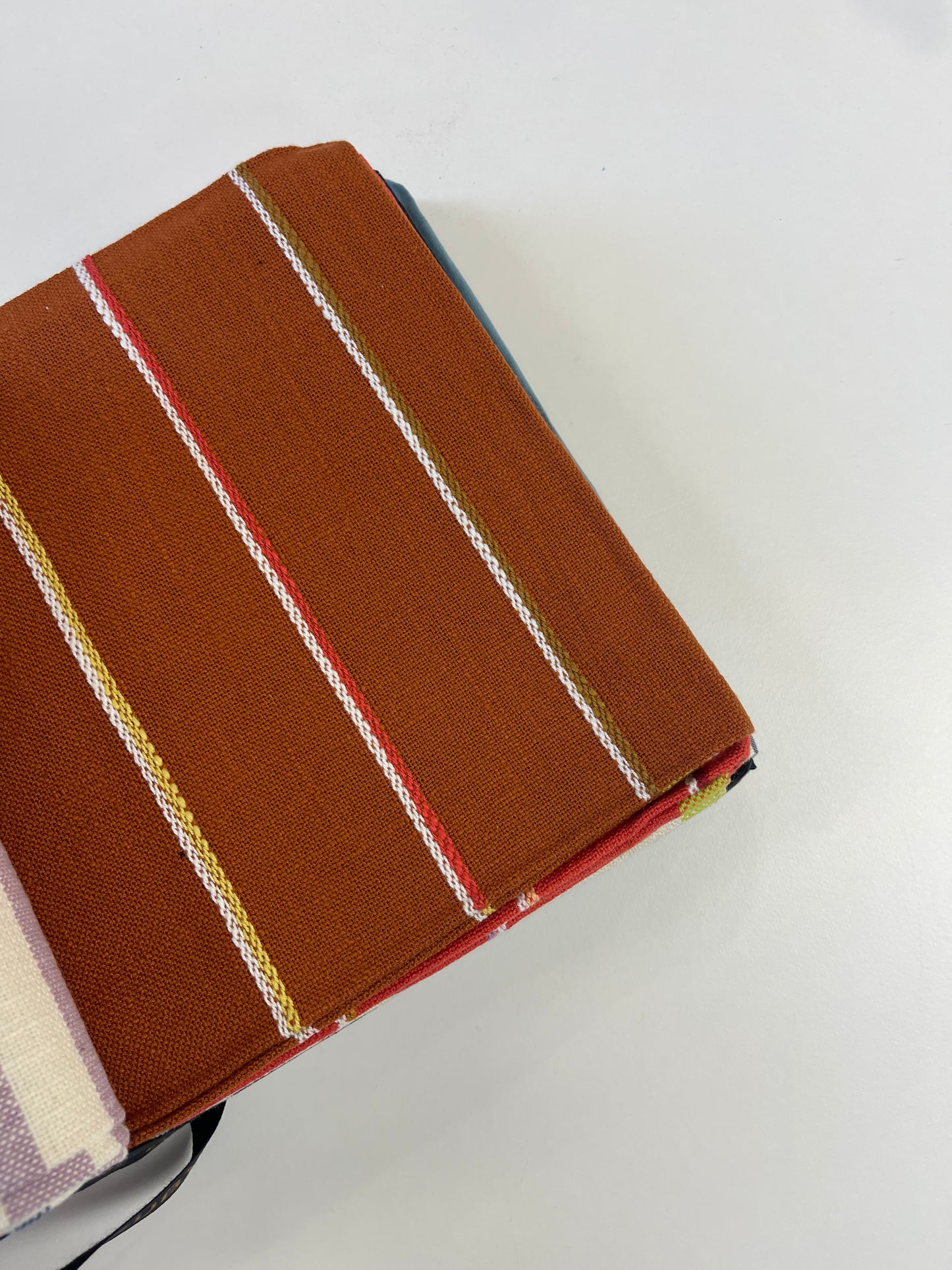 A close-up of a brown woven fabric item, likely a notebook cover or book sleeve, featuring vertical stripes in white, red, yellow, and green, photographed on a plain white surface.