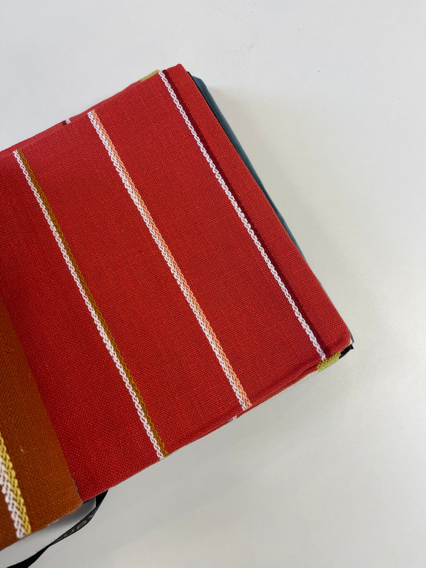 A close-up of a red woven fabric item, likely a notebook cover or book sleeve, featuring vertical stripes in white, gold, and brown, photographed on a plain white surface.