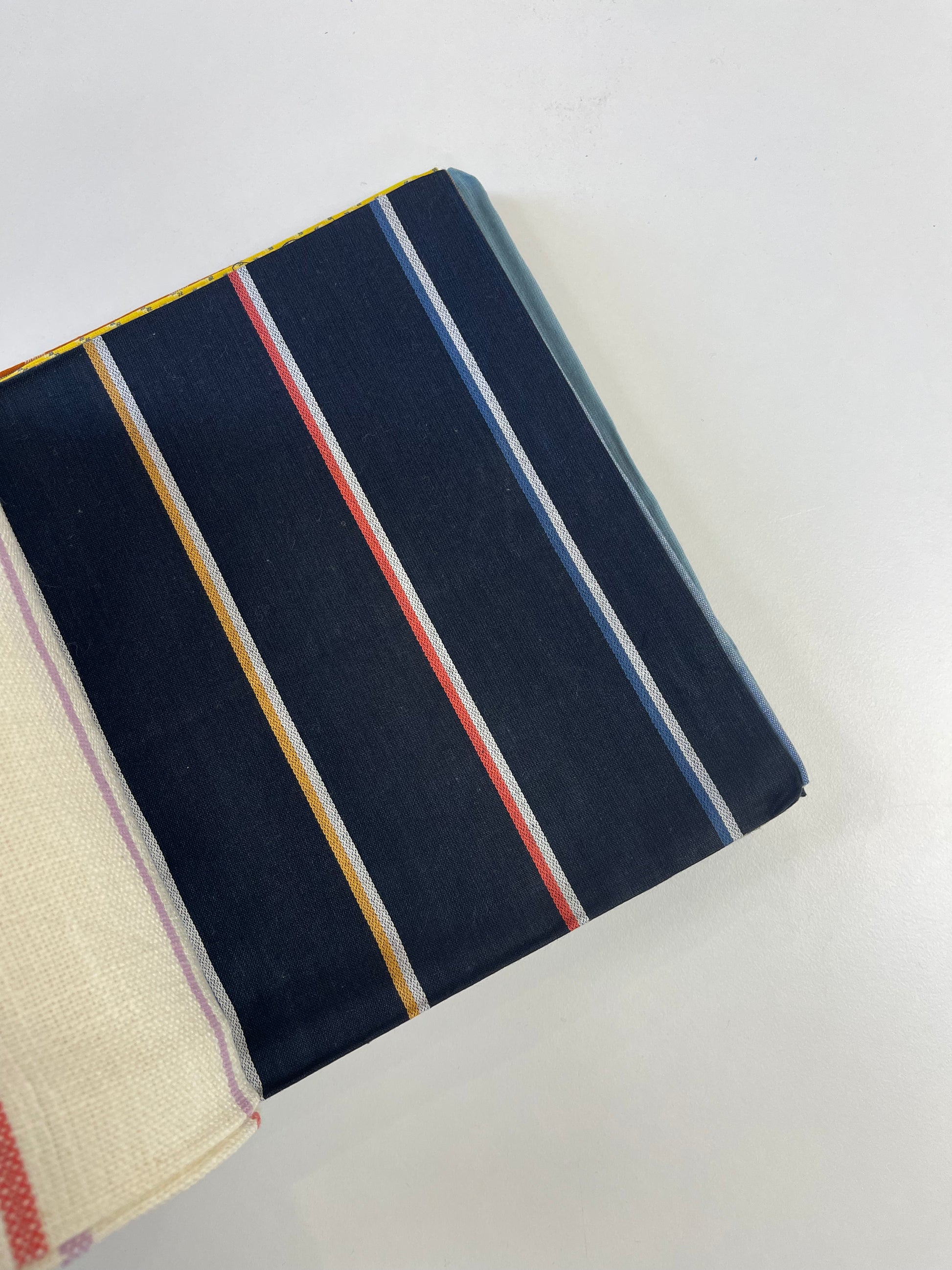 A close-up of a striped fabric product, likely a notebook cover or pouch, featuring a navy blue base with vertical stripes in yellow, red, light blue, and white. A cream-colored woven fabric strip is visible on the left edge.