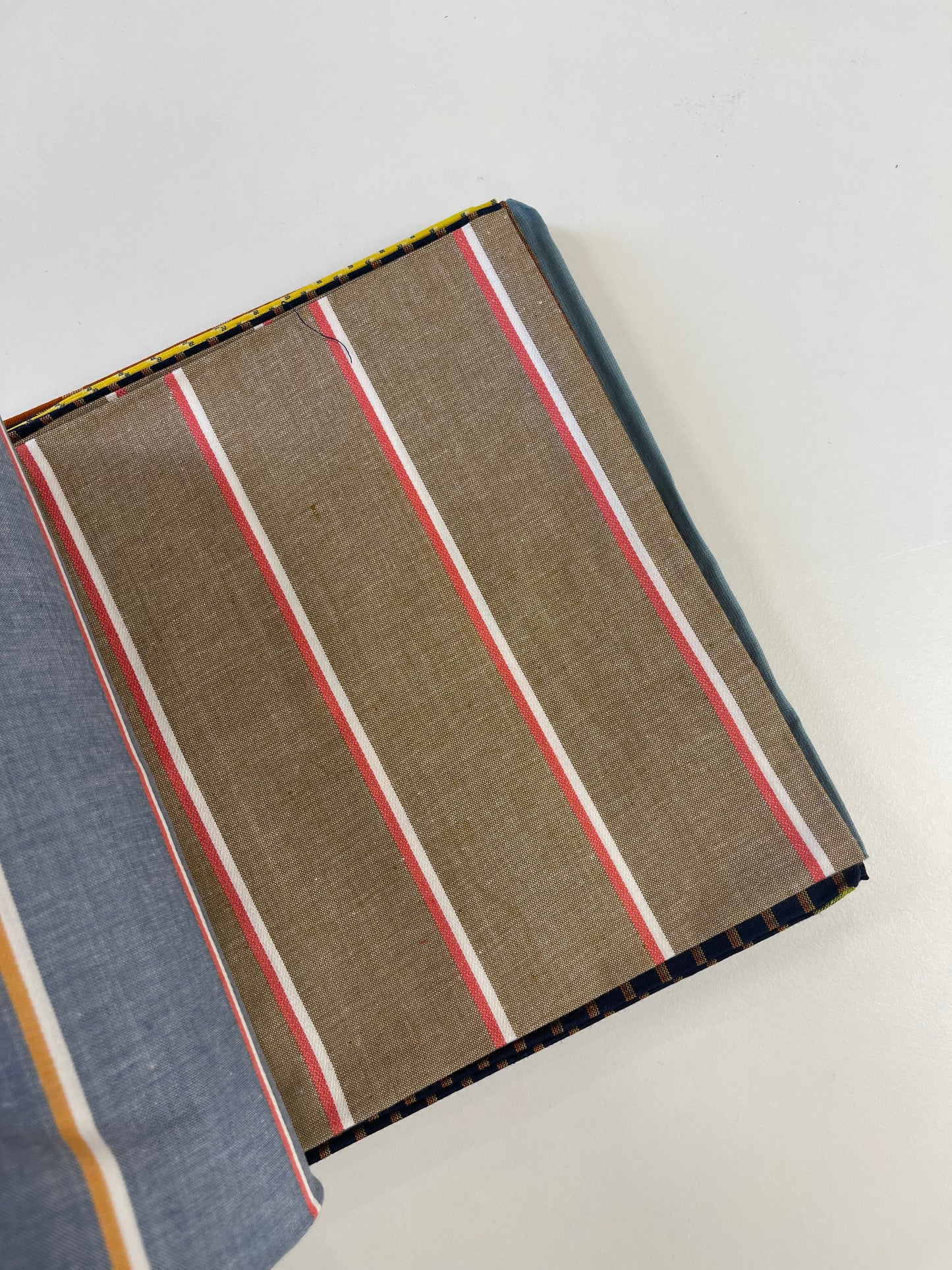 A close-up of a striped fabric or textile sample laid on a white background, showing a brown base with vertical red, white, and thin yellow stripes, plus a blue-gray section along the left edge.