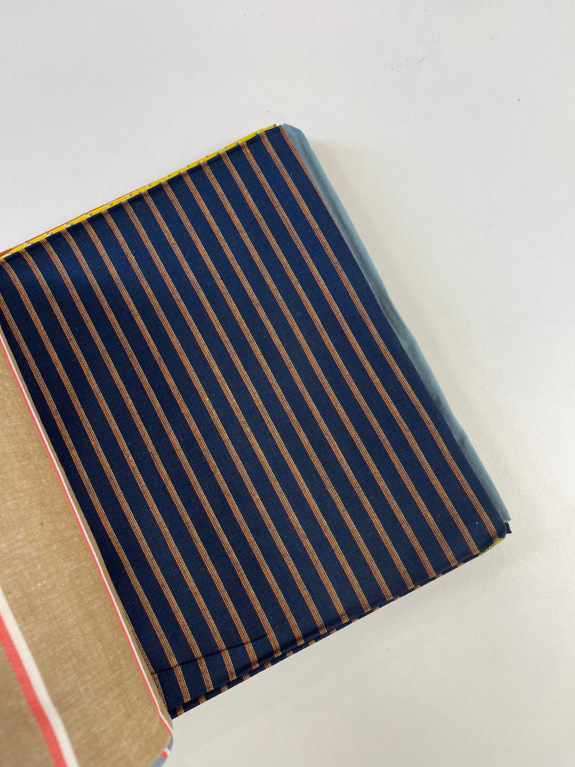 A close-up of a folded fabric item, likely a wallet or pouch, featuring a navy blue background with vertical tan stripes, shown on a plain light background.