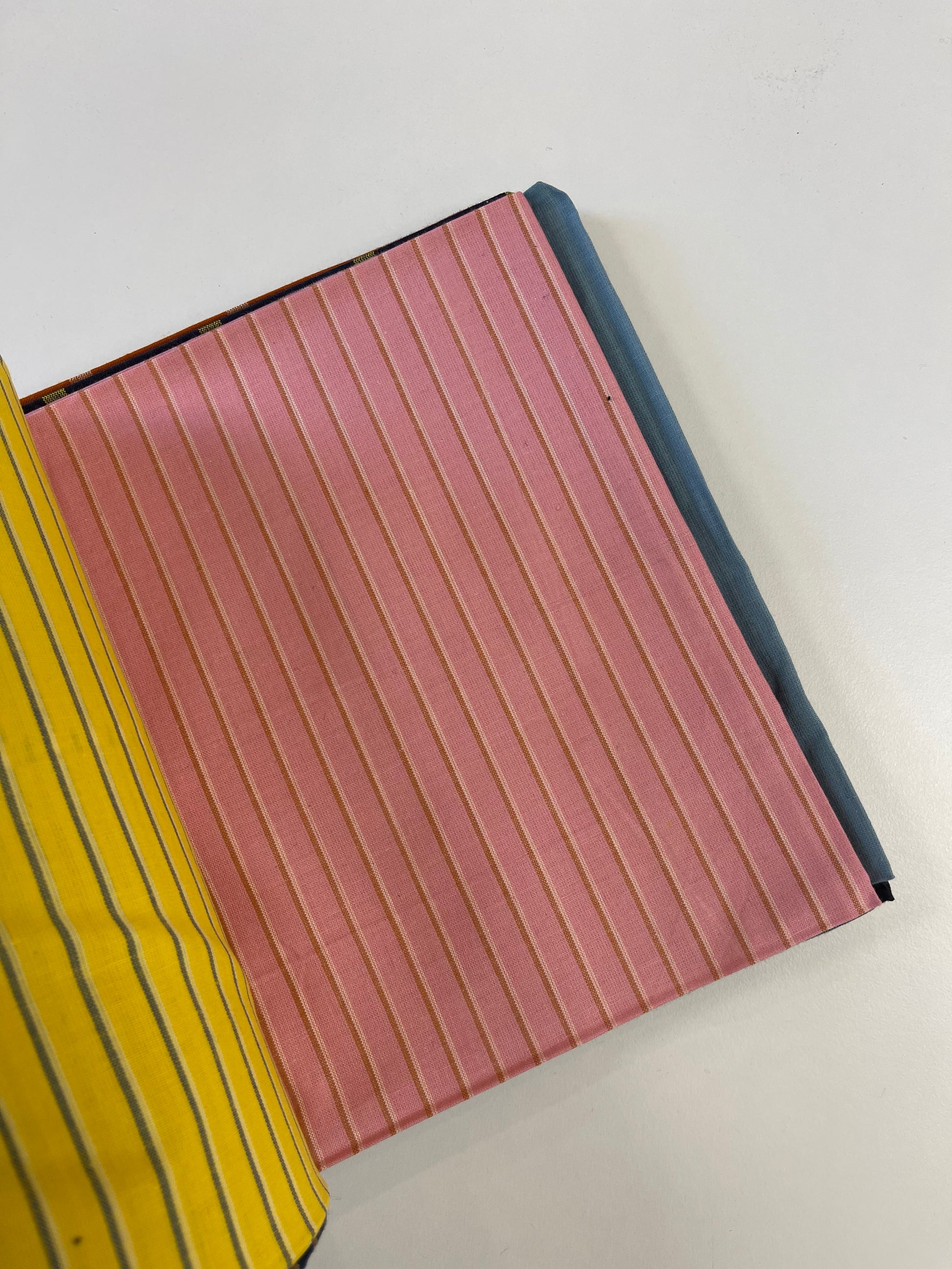 A close-up of a pink striped fabric or material sample with vertical lines, partially overlapped by a yellow striped fabric on the left, on a plain white background.
