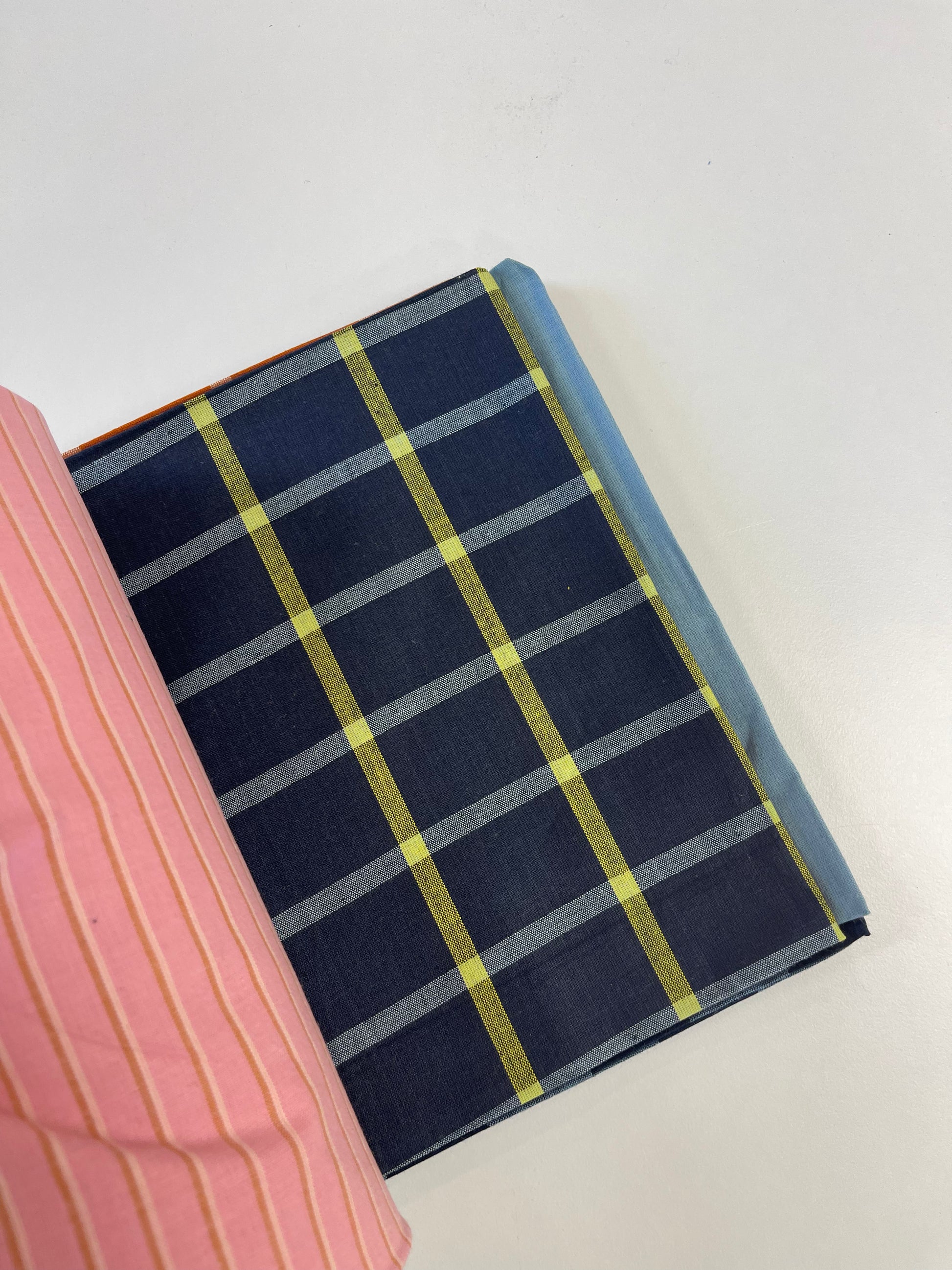A close-up of a navy blue fabric item, likely a notebook cover or book sleeve, featuring a yellow and gray plaid pattern, with a pink striped fabric partially visible on the left.