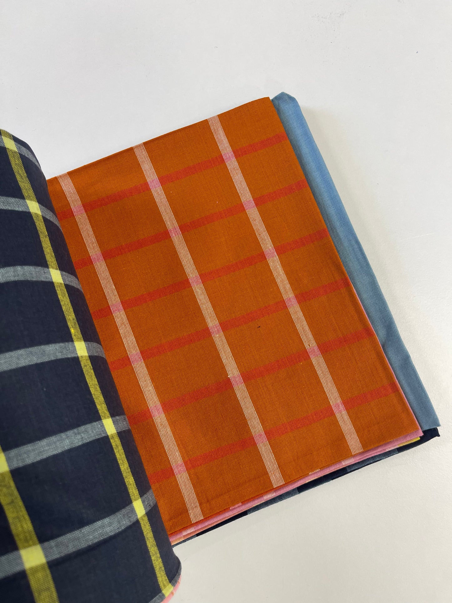 A folded fabric sample or book showing textile colors and patterns, with a prominent orange page featuring thin pink and cream stripes, a navy page with yellow and gray plaid on the left, and a light blue page on the right, photographed on a white background.