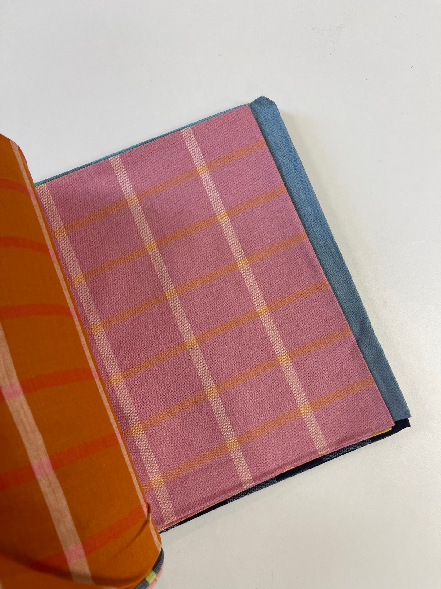 A folded piece of pink plaid fabric with orange and white stripes, layered over a blue fabric, with a folded orange striped fabric visible on the left, set against a plain white background.