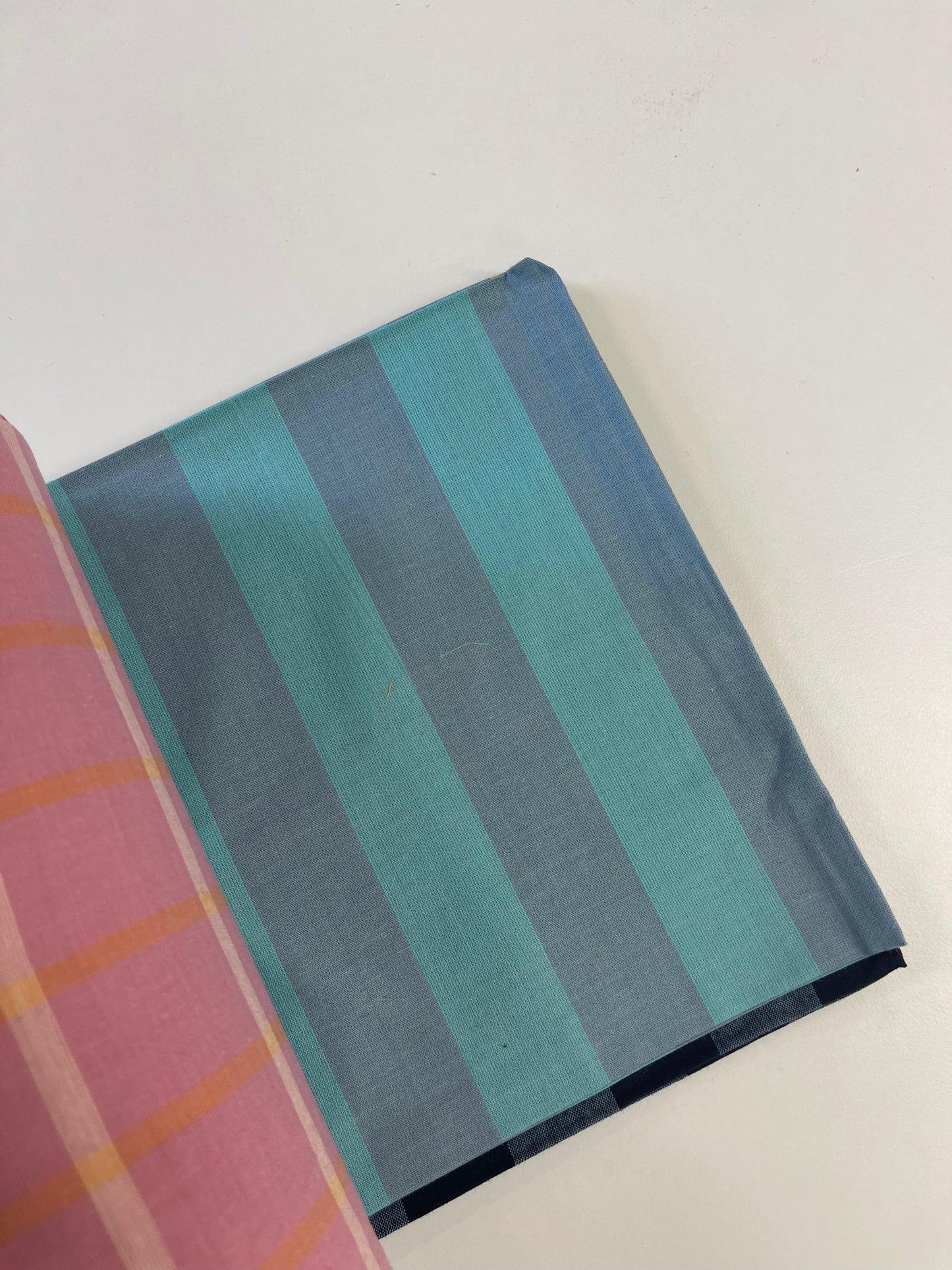 A folded piece of fabric with vertical stripes in teal, blue, and gray, partially overlapping with a pink striped fabric on a plain white surface.
