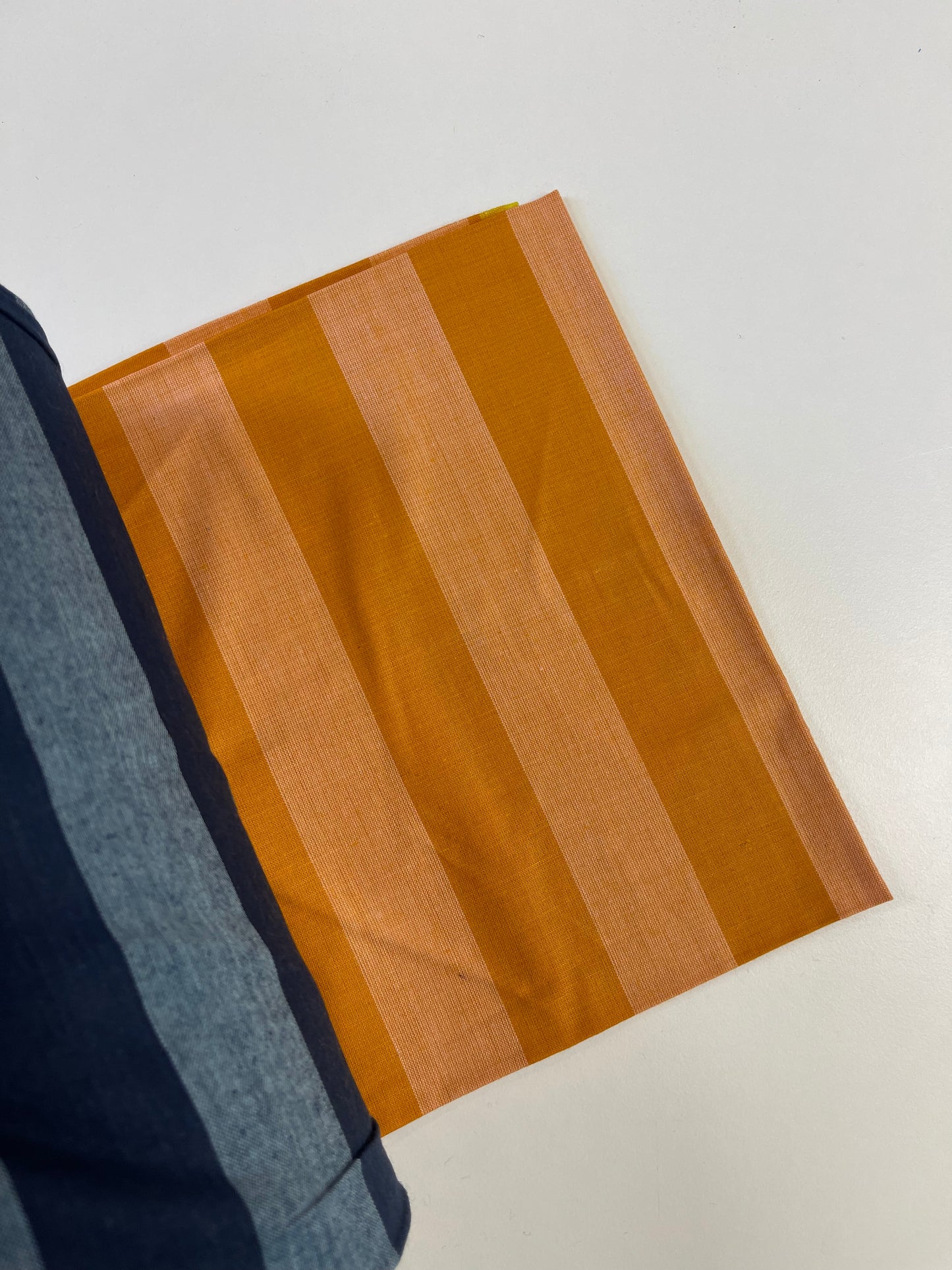 A close-up of a striped fabric swatch with alternating bands of mustard yellow and light peach, shown alongside a folded navy blue striped textile on a plain white surface.