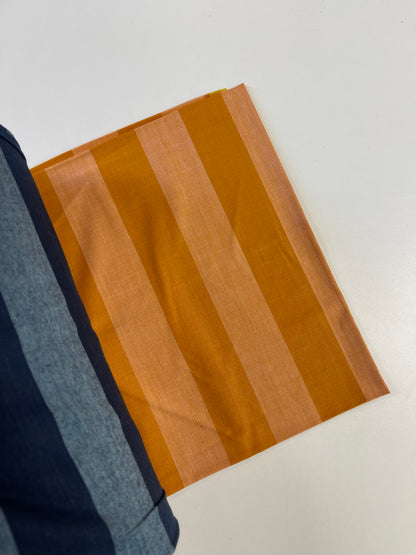 A close-up of a striped fabric swatch with alternating bands of mustard yellow and light peach, shown alongside a folded navy blue striped textile on a plain white surface.