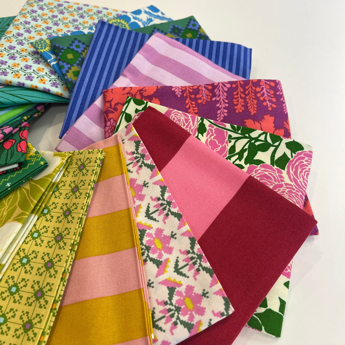 A collection of colorful patterned fat quarter fabric squares arranged in a circular stack on a plain white background. Form Anna Maria Textiles for quilting and sewing. 