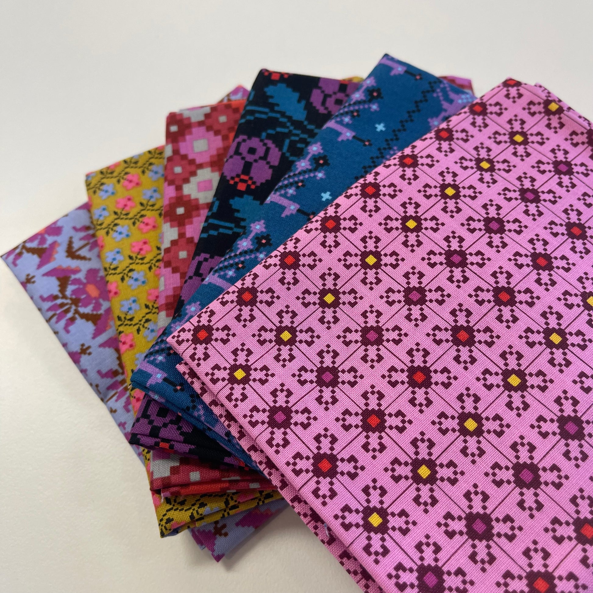 A stack of folded patterned fabric pieces fanned out on a white surface, showing multiple colorful designs including pink, blue, yellow, and purple geometric and floral motifs.