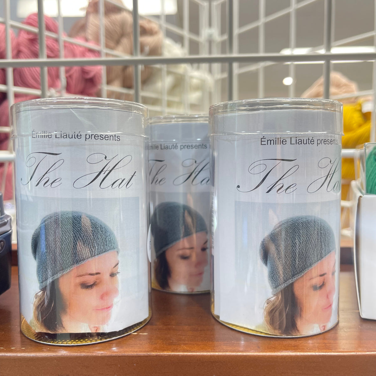 Three clear cylindrical jars labeled 'The Hat' by Émilie Liauté, featuring a photo of a woman wearing a gray knit hat on the front, displayed on a wooden surface.