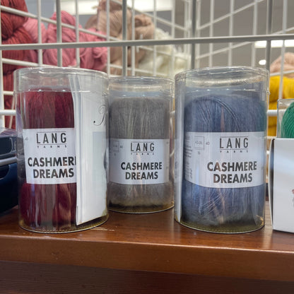 Three clear plastic jars of Lang Yarns 'Cashmere Dreams' yarn in burgundy, taupe, and slate blue, displayed on a wooden shelf in a store.