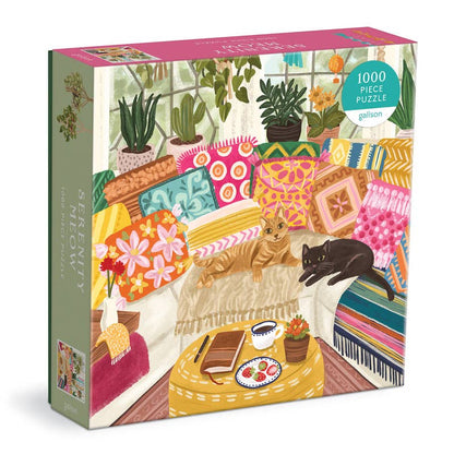 A boxed jigsaw puzzle package shown at a slight angle on a white background. The front artwork depicts a cozy living room scene with colorful patterned cushions, plants, a coffee table, and two cats, one orange and one black.