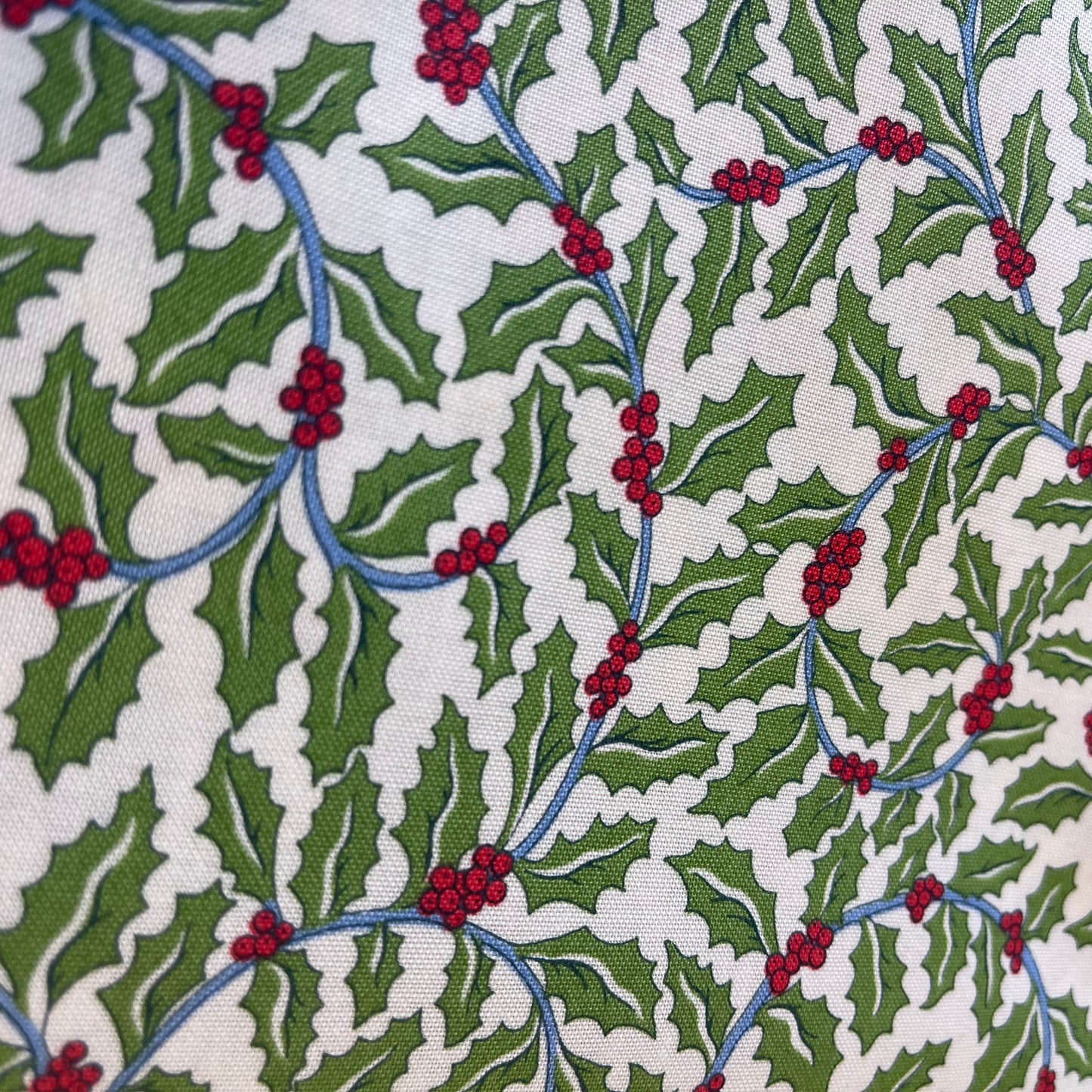 A close-up of patterned fabric with a repeating holly design featuring green leaves, red berries, and blue stems on an off-white background from Liberty of London Fabrics for quilting and crafting.