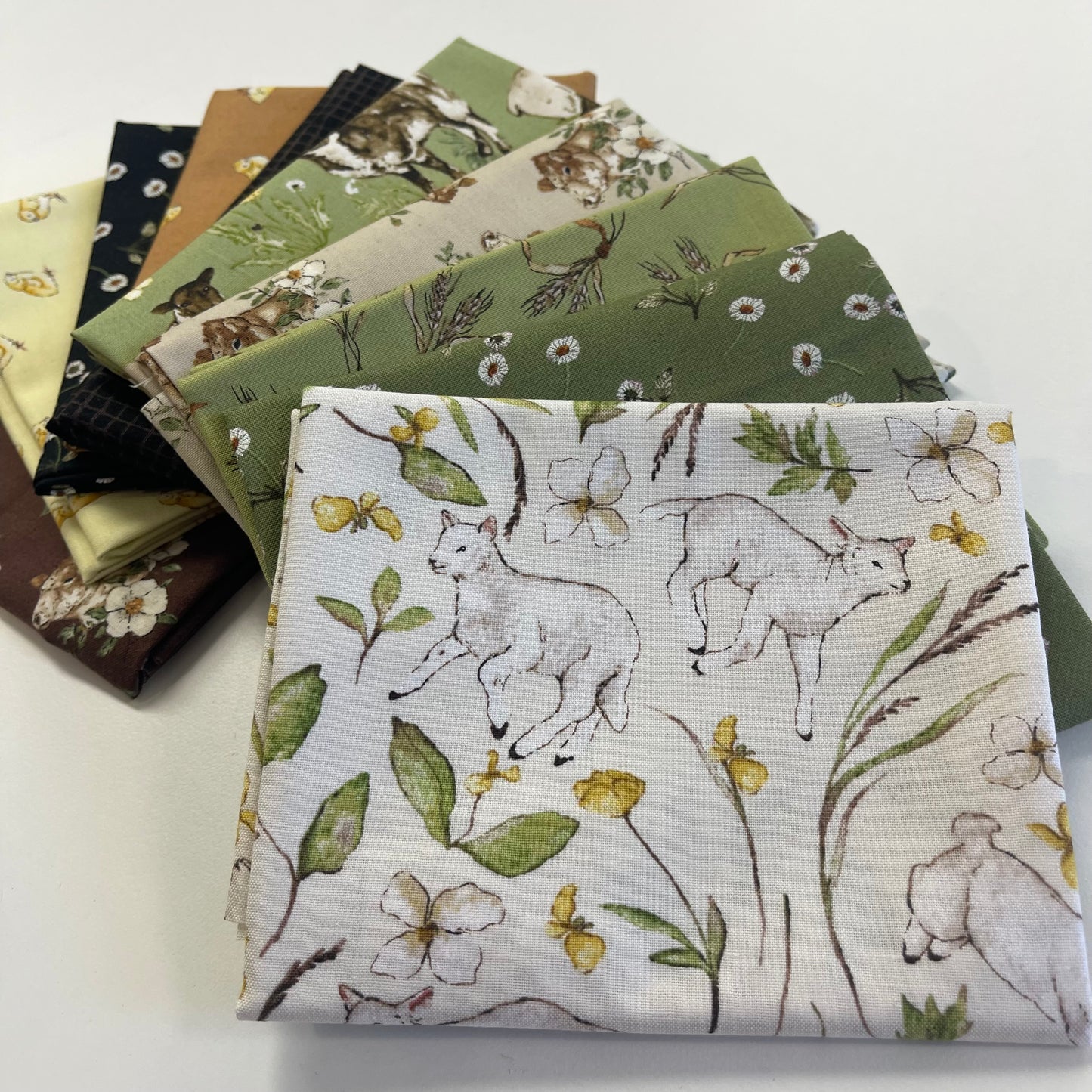 A stack of fabric swatches fanned out on a white surface, with the top fabric featuring a print of white rabbits, green leaves, and yellow wildflowers on an off-white background.