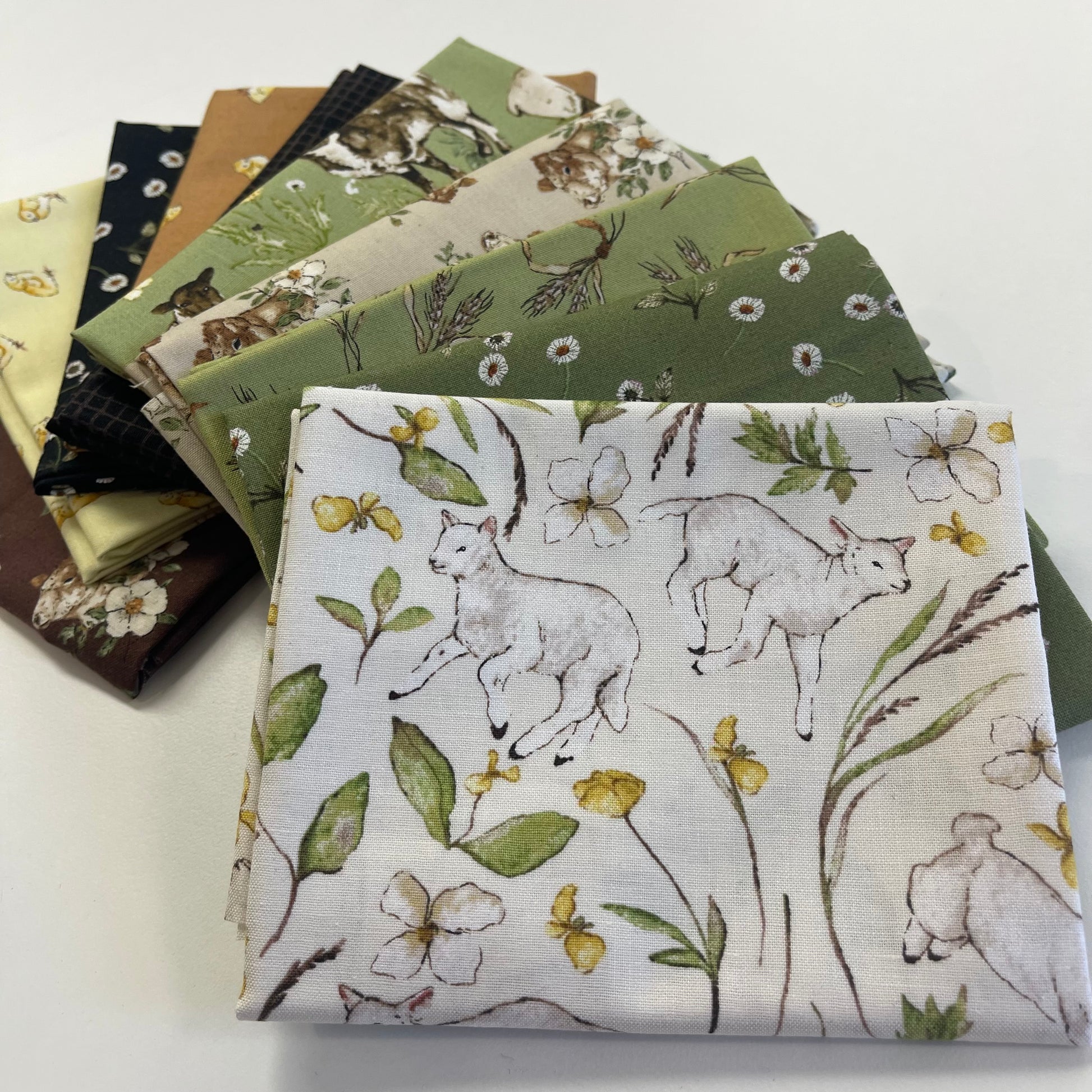 A stack of fabric swatches fanned out on a white surface, with the top fabric featuring a print of white rabbits, green leaves, and yellow wildflowers on an off-white background.