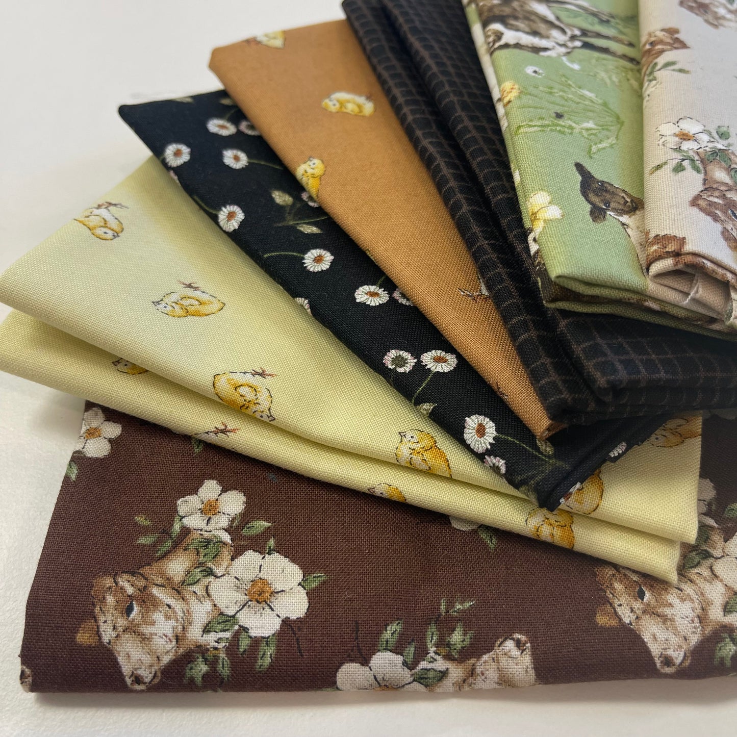 A stack of folded fabric samples with various spring and nature-themed prints, including a brown bear, white flowers, yellow chicks, daisies, a bird, and green foliage, displayed on a light neutral background.