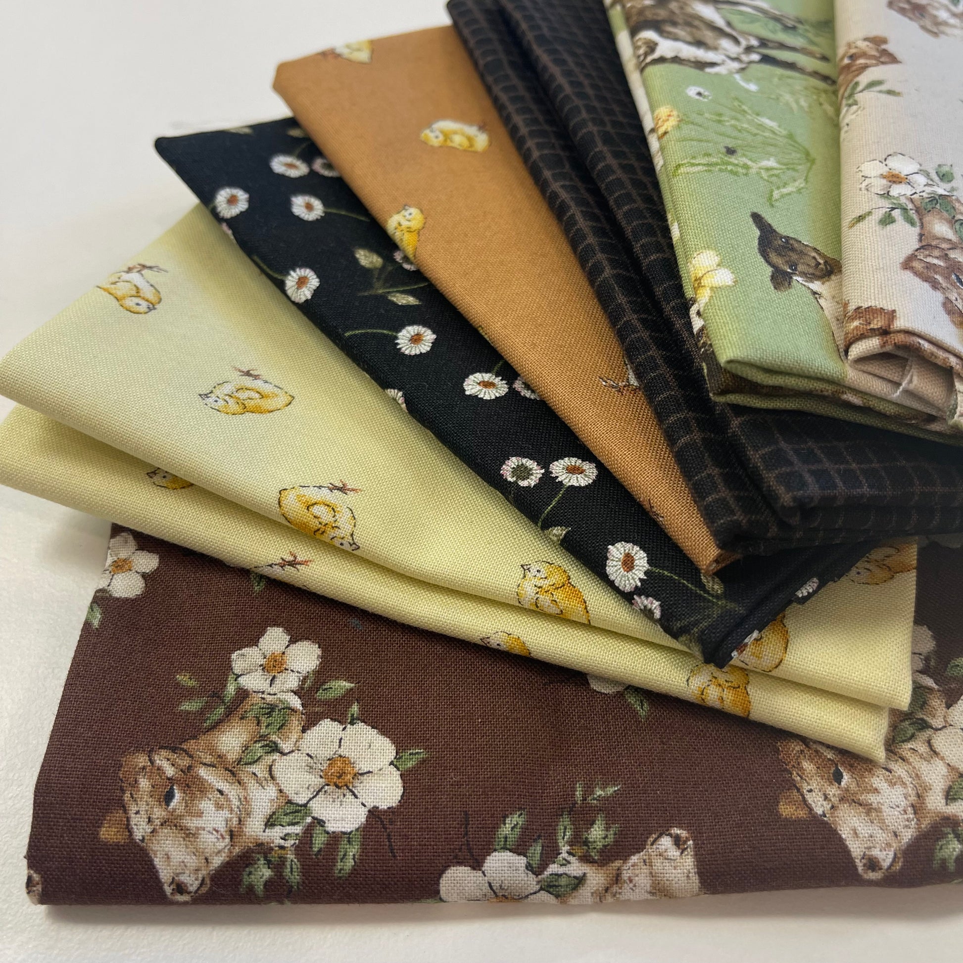 A stack of folded fabric samples with various spring and nature-themed prints, including a brown bear, white flowers, yellow chicks, daisies, a bird, and green foliage, displayed on a light neutral background.