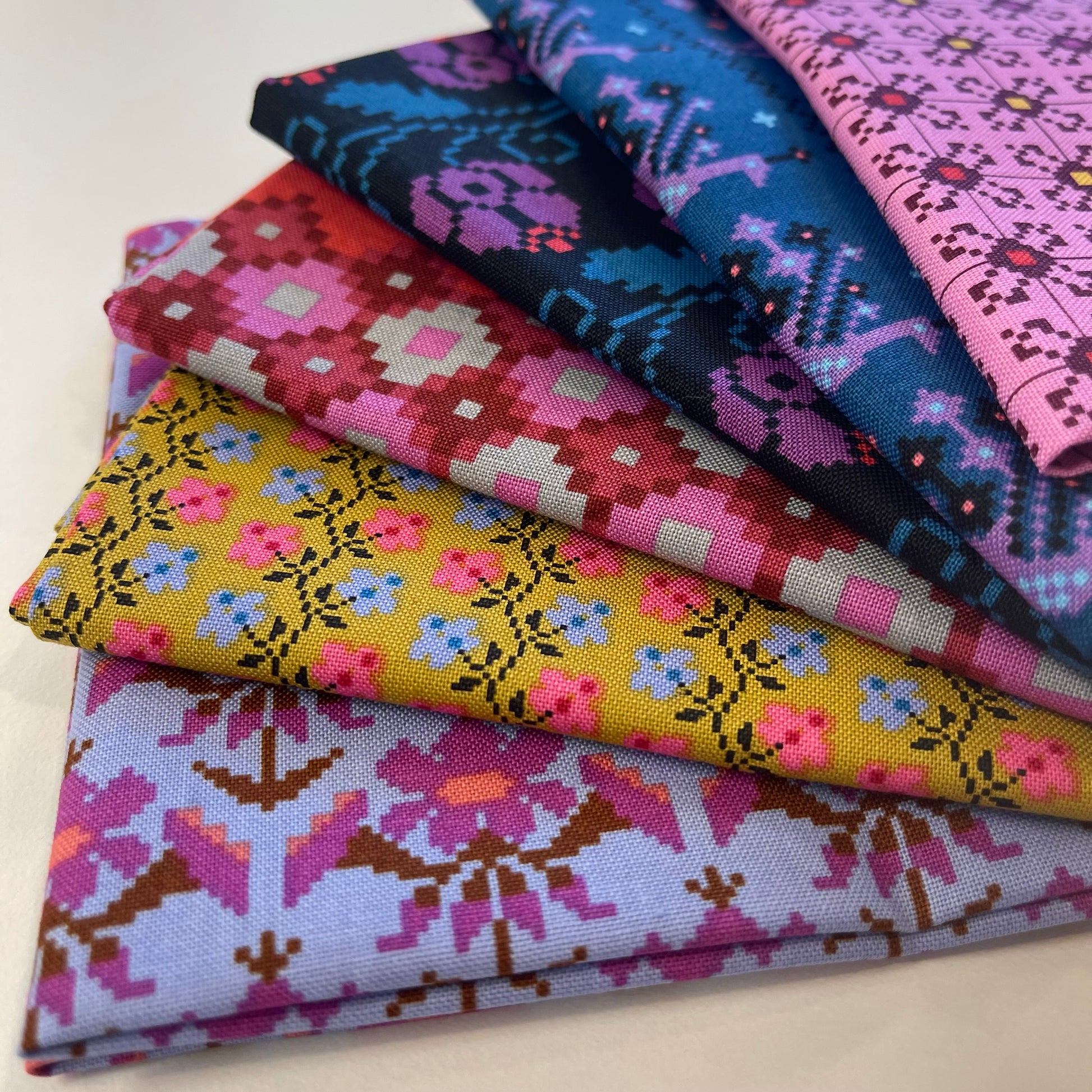 A stack of folded patterned fabric pieces in multiple colors, including pink, blue, yellow, purple, and red geometric and floral motifs, photographed on a light neutral background.