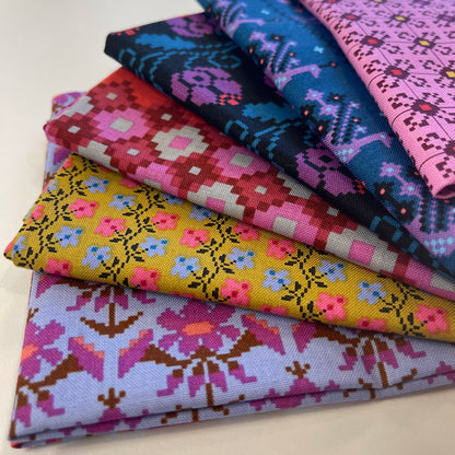 A stack of folded patterned fabric pieces in multiple colors, including pink, blue, yellow, purple, and red geometric and floral motifs, photographed on a light neutral background.