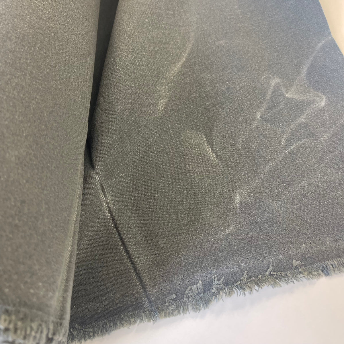 A roll of dark medium waxed canvas fabric with a frayed edge, partially unrolled on a plain white background.