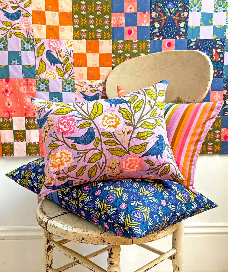 Colorful pillows with floral and bird patterns on a chair in front of a patchwork wall.