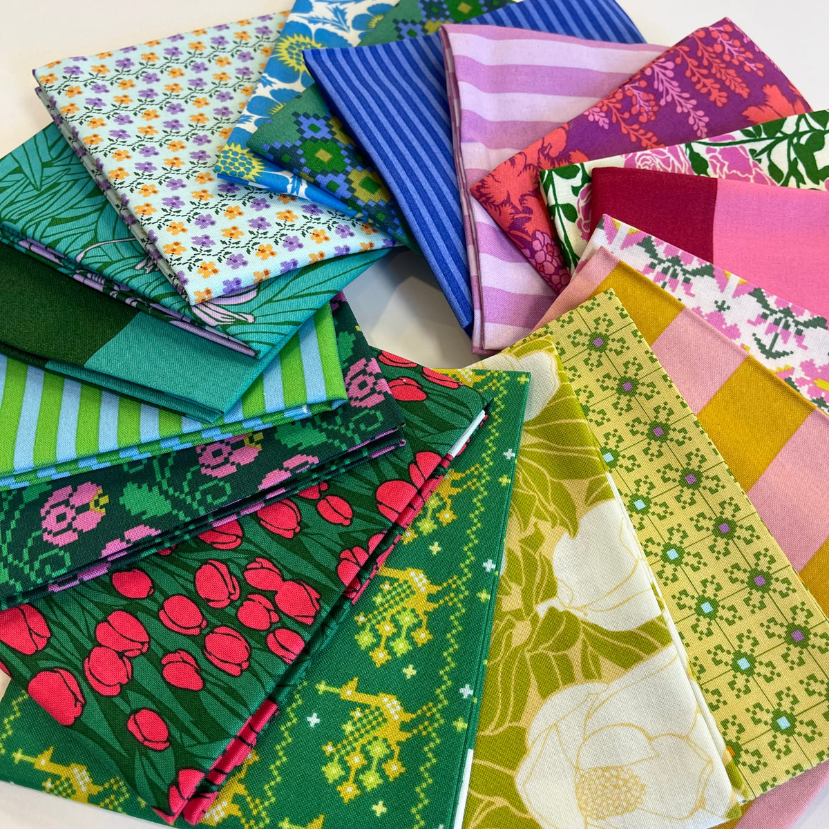 A collection of colorful patterned fat quarter fabric squares arranged in a circular stack on a plain white background. Form Anna Maria Textiles for quilting and sewing. 