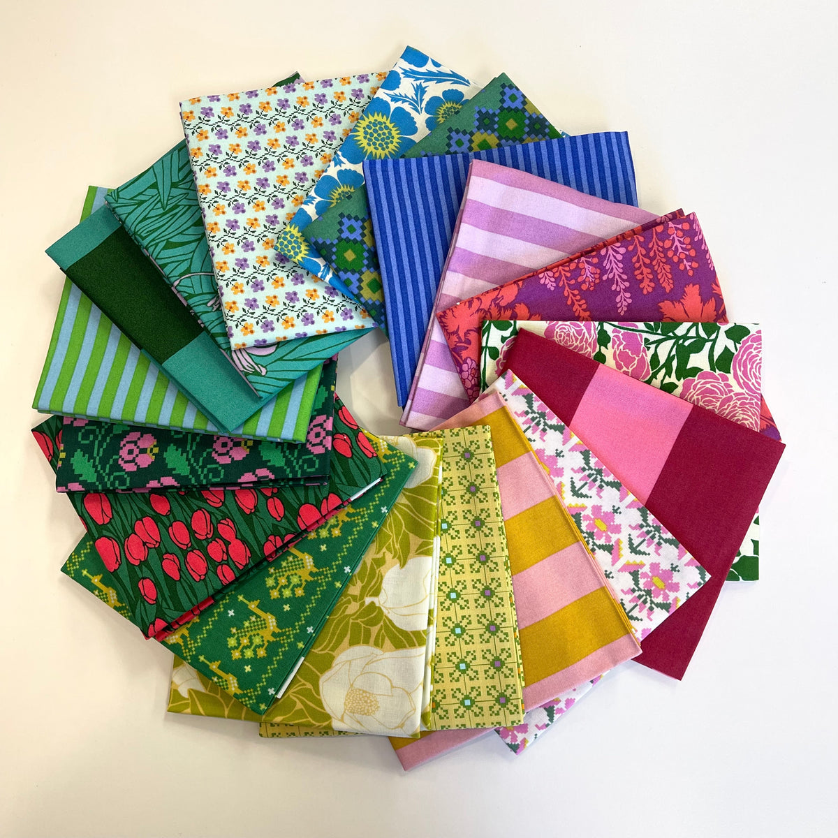 A collection of colorful patterned fat quarter fabric squares arranged in a circular stack on a plain white background. Form Anna Maria Textiles for quilting and sewing. 