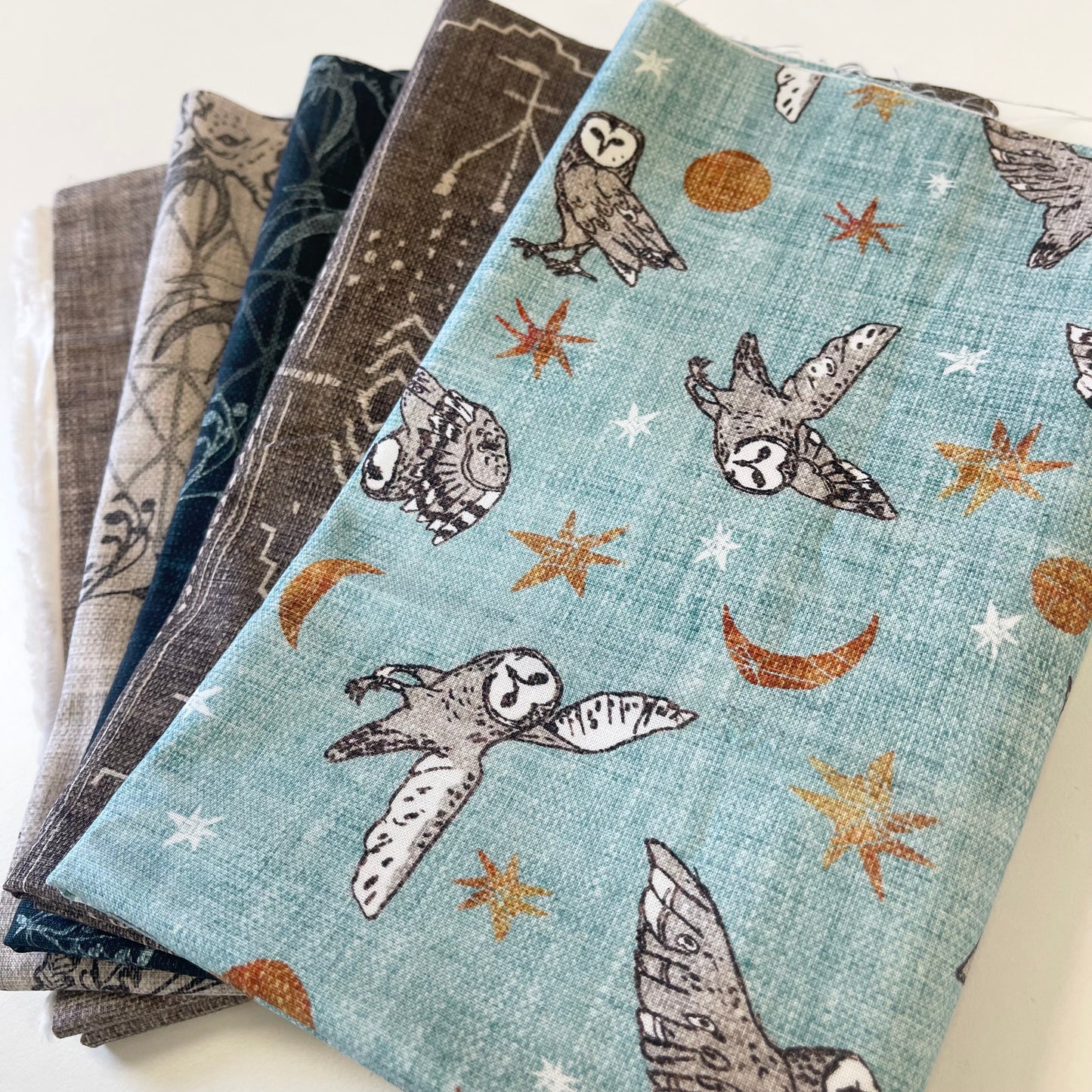 A stack of folded patterned fabrics on a white surface, with the top fabric featuring a light blue background printed with owls, crescent moons, and star-like shapes in gray, white, and rust orange.