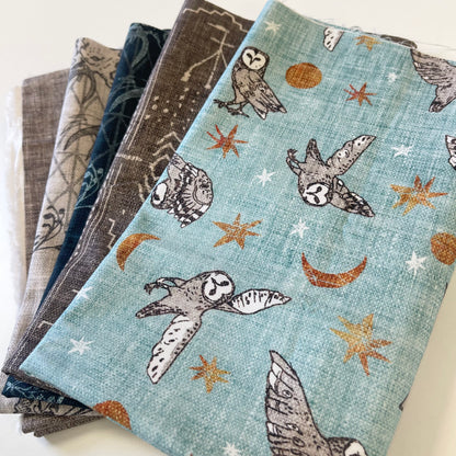 A stack of folded patterned fabrics on a white surface, with the top fabric featuring a light blue background printed with owls, crescent moons, and star-like shapes in gray, white, and rust orange.