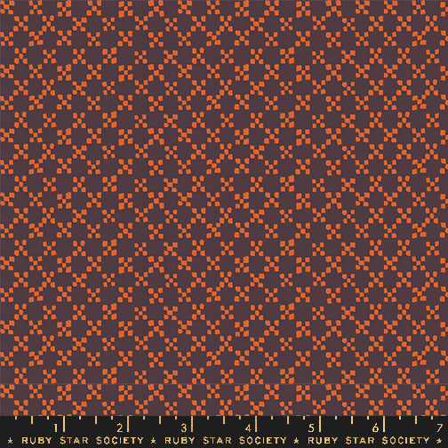 A close-up fabric pattern swatch showing a repeating geometric design in rust orange and dark plum purple, with a Ruby Star Society ruler along the bottom edge for scale.