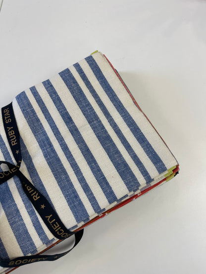 A folded stack of striped fabric napkins or table linens tied with a black ribbon printed with 'RIPY STARS SOCIETY', showing blue and white stripes on top, photographed on a white surface.