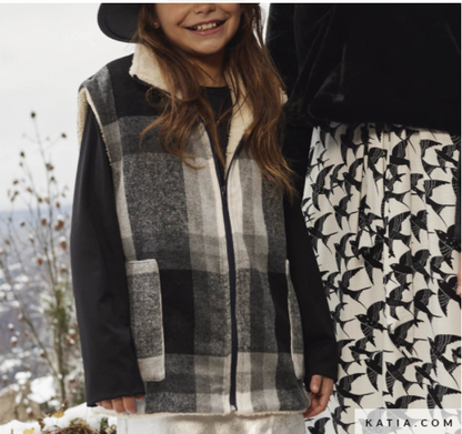 A child wearing a black and gray plaid zip-front vest with a cream sherpa collar, standing outdoors next to another person in a black-and-white bird-print skirt.