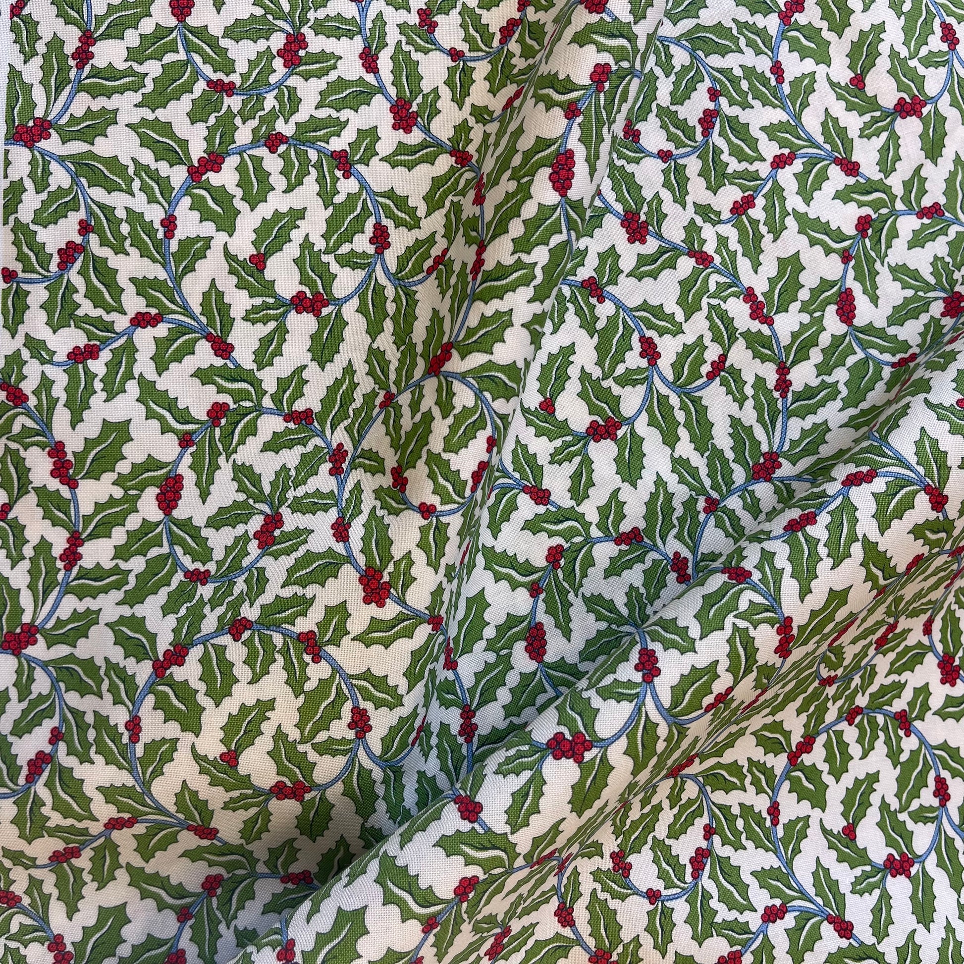 A close-up of patterned fabric with a repeating holly design featuring green leaves, red berries, and blue stems on an off-white background from Liberty of London Fabrics for quilting and crafting.