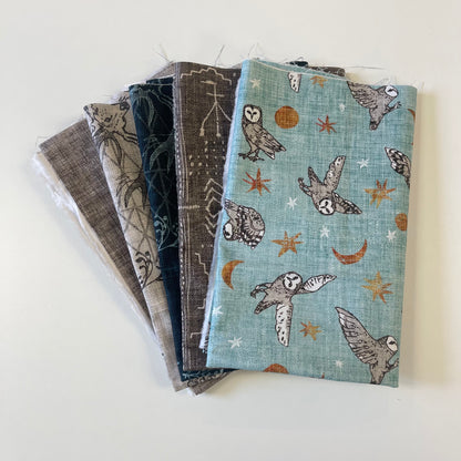 A stack of folded fabric pieces with various patterns, including a light blue one featuring owls, stars, and crescent moons, arranged on a plain white surface.