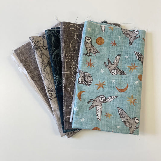 A stack of folded fabric pieces with various patterns, including a light blue one featuring owls, stars, and crescent moons, arranged on a plain white surface.