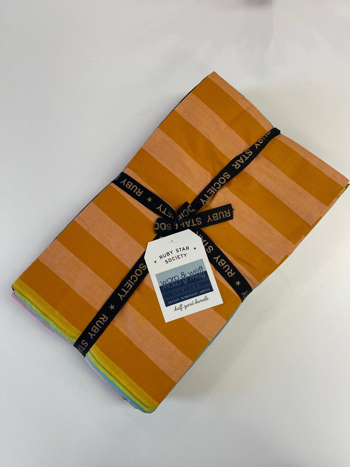 A folded stack of Ruby Star Society woven cotton fabrics with orange and tan stripes, wrapped with a black branded ribbon and a white paper tag, on a white background.