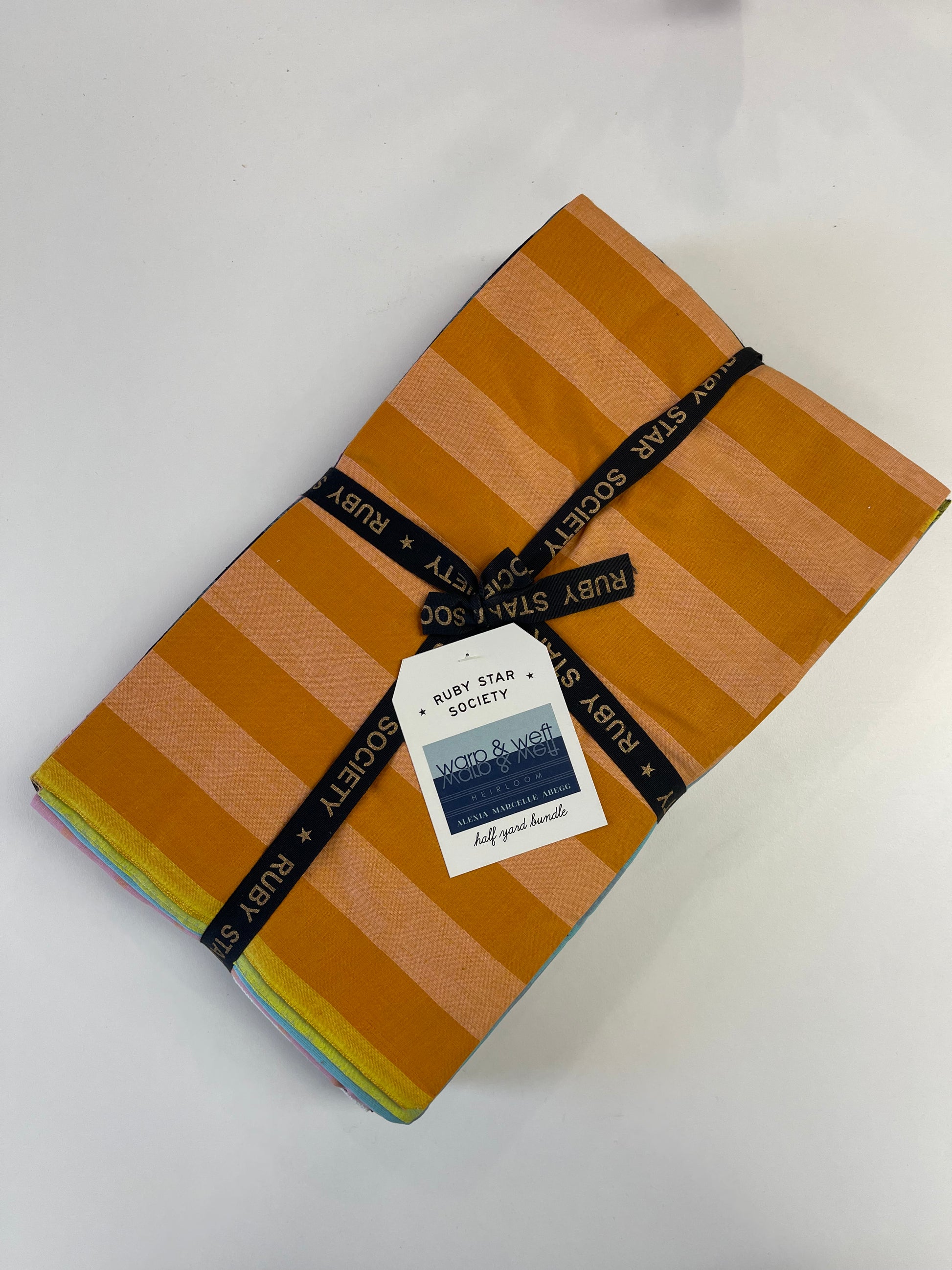 A folded stack of Ruby Star Society woven cotton fabrics with orange and tan stripes, wrapped with a black branded ribbon and a white paper tag, on a white background.