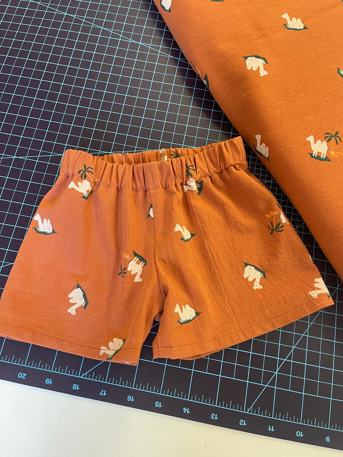A close-up of rust-orange fabric shorts with a white and green llama print, laid flat on a black gridded cutting mat next to a matching rolled fabric piece.