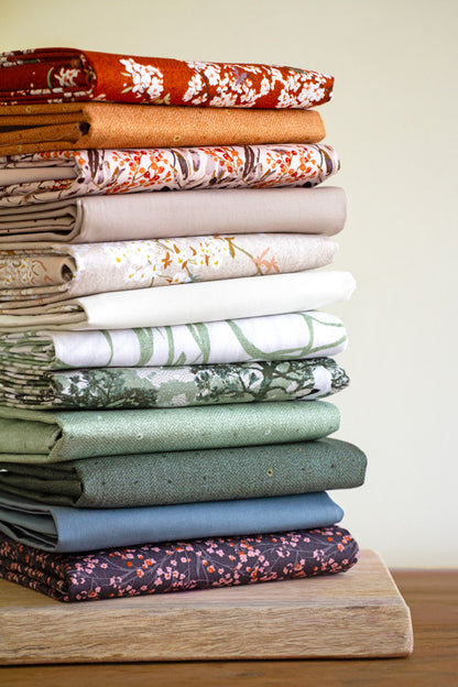 Stack of folded fabric swatches in various colors on a wooden surface from Soar by Katerina Roccella Art Gallery Fabrics premium cotton for sewing and crafting.