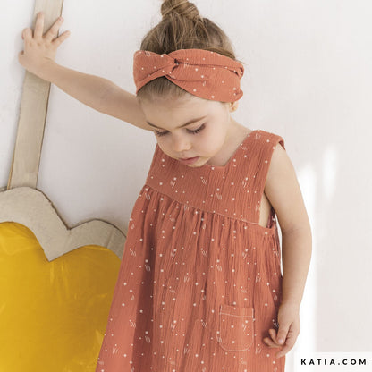 Young girl wearing a coral colored dress with a matching headband, standing against a white wall.