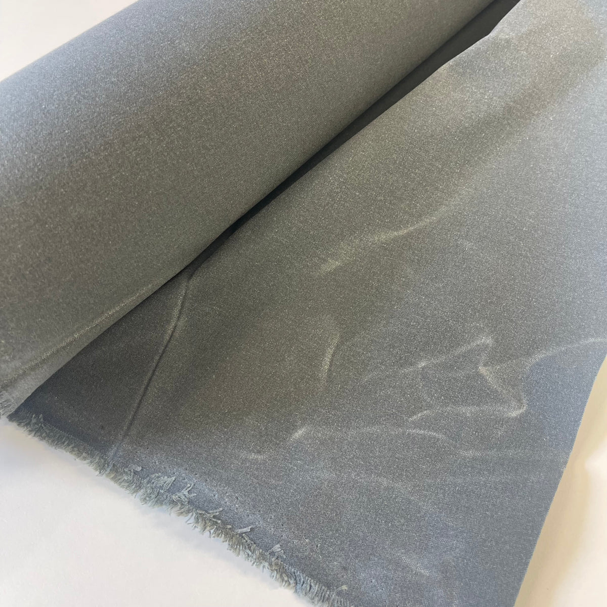 A roll of dark medium waxed canvas fabric with a frayed edge, partially unrolled on a plain white background.